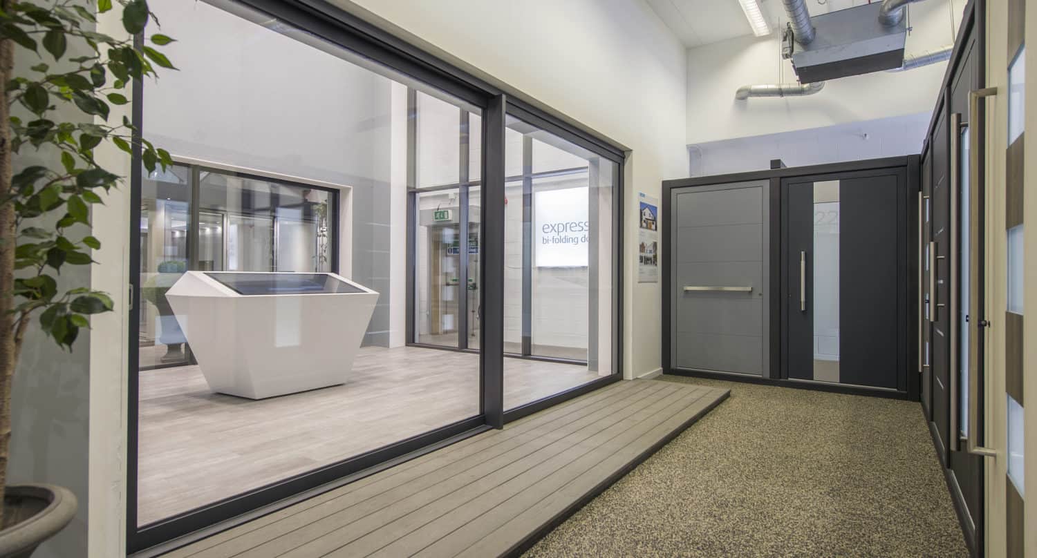 A modern showroom interior with large sliding doors and windows, a gray front door display, a touchscreen kiosk, bright lighting, and a potted plant on the left.
