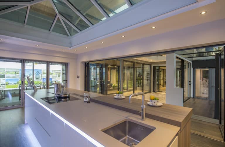 A modern kitchen with a sleek island, built-in sink, and stovetop sits beneath a large glass ceiling. Bespoke glass solutions like sliding doors and windows flood the open, contemporary living space with natural light.
