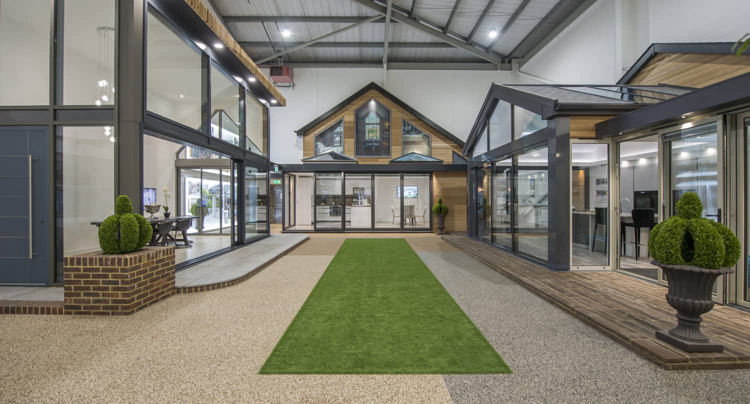A modern indoor showroom displays several bespoke glass solutions, including glass-walled home extensions and conservatories, with a green artificial grass walkway and decorative potted plants under a high industrial ceiling.