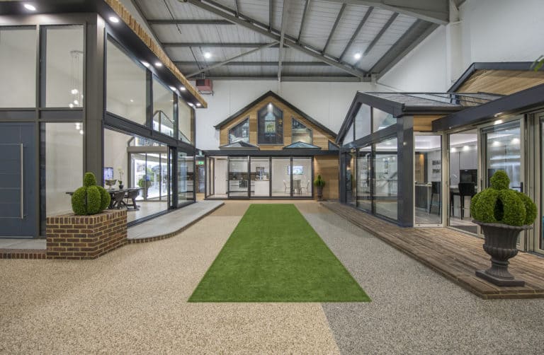 A modern indoor showroom displays several bespoke glass solutions, including glass-walled home extensions and conservatories, with a green artificial grass walkway and decorative potted plants under a high industrial ceiling.