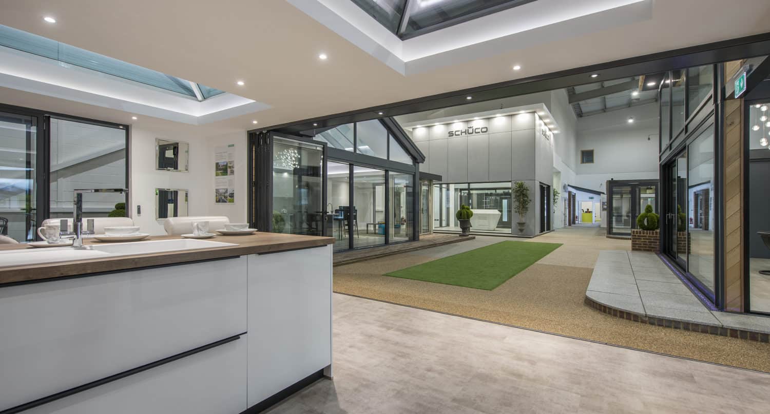 Modern indoor showroom with large glass windows, skylights, and contemporary fixtures. The space features a green carpet strip, potted plants, and displays of front doors and windows under bright lighting.