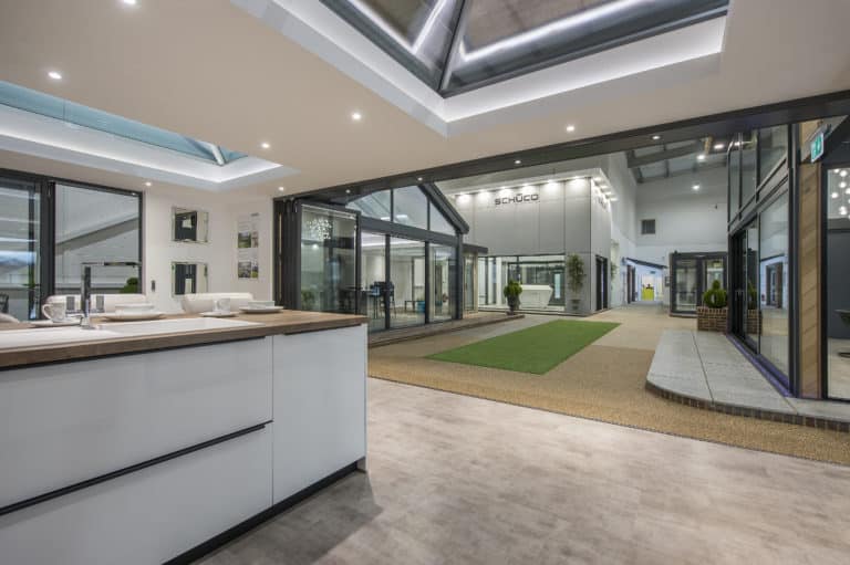 Modern indoor showroom with large glass windows, skylights, and contemporary fixtures. The space features a green carpet strip, potted plants, and displays of front doors and windows under bright lighting.