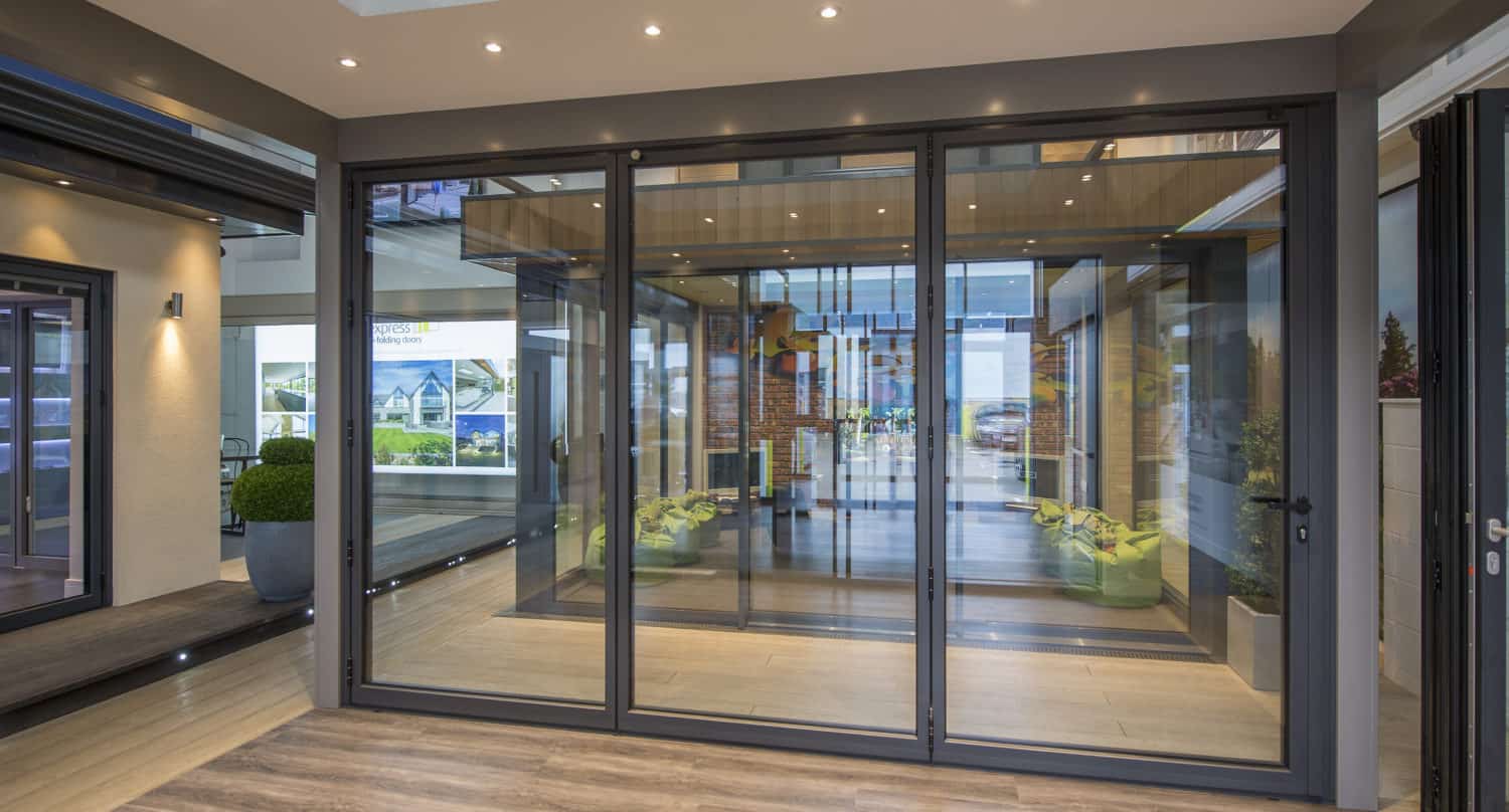 A modern room with large sliding doors, glass roofing, recessed ceiling lights, wood flooring, and indoor plants visible through the panels creates a bright and open atmosphere.