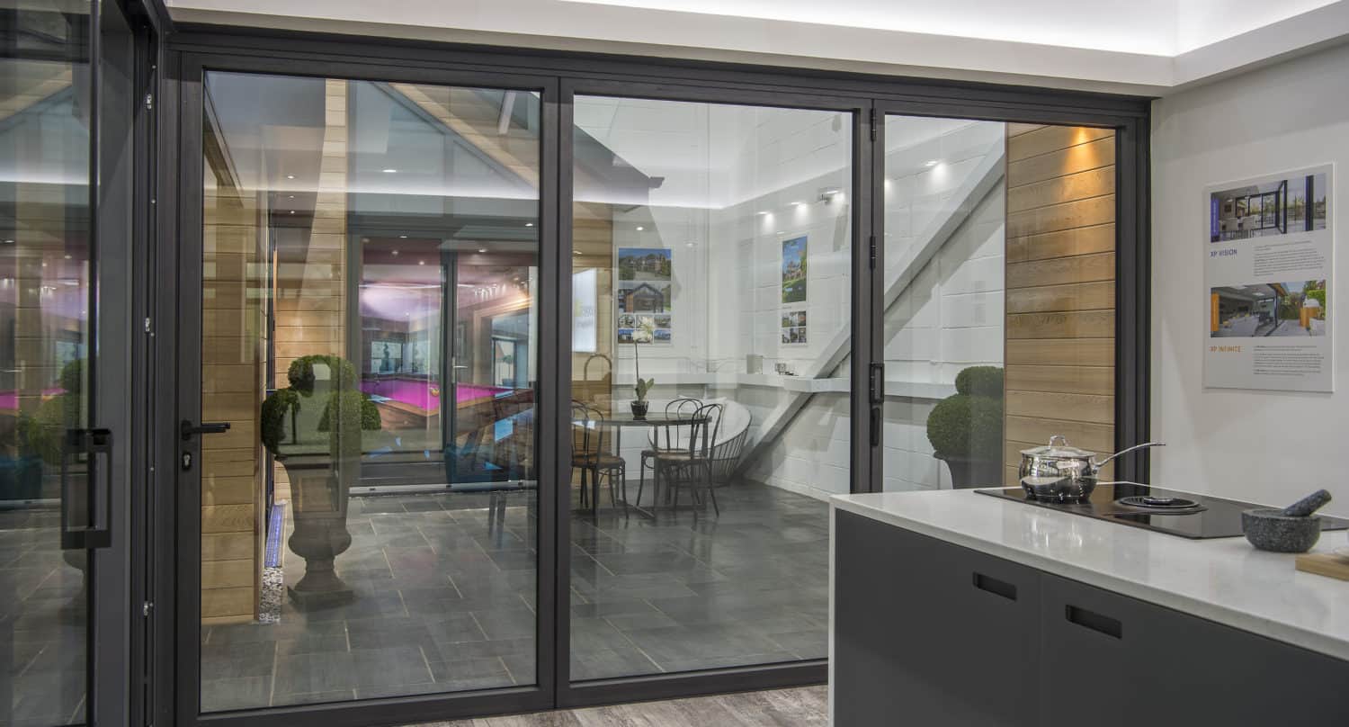 Modern kitchen with sleek grey cabinets and a cooktop, separated by large bifold doors from a stylish dining area with metal chairs and wall art, featuring contemporary lighting, indoor plants, and bespoke glass solutions.