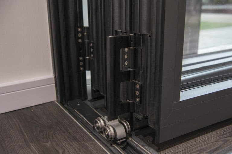 Close-up of the bottom track and hinge mechanism of a black-framed bifold door, showing rollers and metal hardware on a dark wood floor.