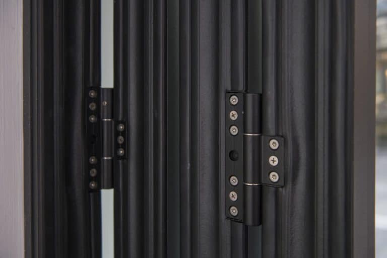 Close-up of metal hinges on black bifold doors, showing the screws and detailed mechanism connecting the door panels. Natural light is visible in the background, highlighting premium bespoke glass solutions.