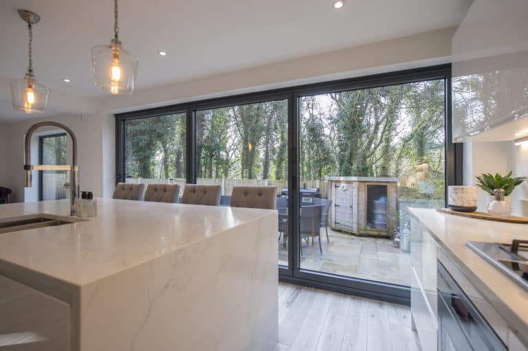 Modern kitchen with a marble island, pendant lights, and bar stools. Expansive sliding doors lead to a patio with outdoor seating, while bespoke glass solutions enhance views of the surrounding trees and greenery.