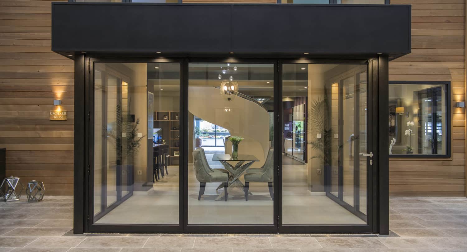 Modern glass-walled patio room with bifold doors, two chairs, and a small round table set for a meal. Wooden exterior walls and large windows surround the space, creating a sleek, contemporary look with bespoke glass solutions.