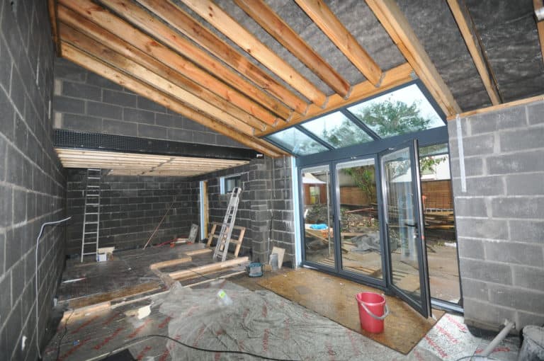 A room under construction with exposed cinder block walls, a sloped wooden ceiling, ladders, building materials, and large sliding doors opening to a backyard. A red bucket sits on the unfinished floor.