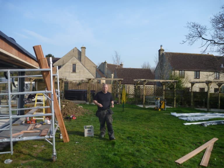 A person stands on a grassy lawn with construction materials and scaffolding nearby. Among scattered beams and tools, bespoke glass solutions for front doors are being prepared, with stone houses and pitched roofs visible in the background.