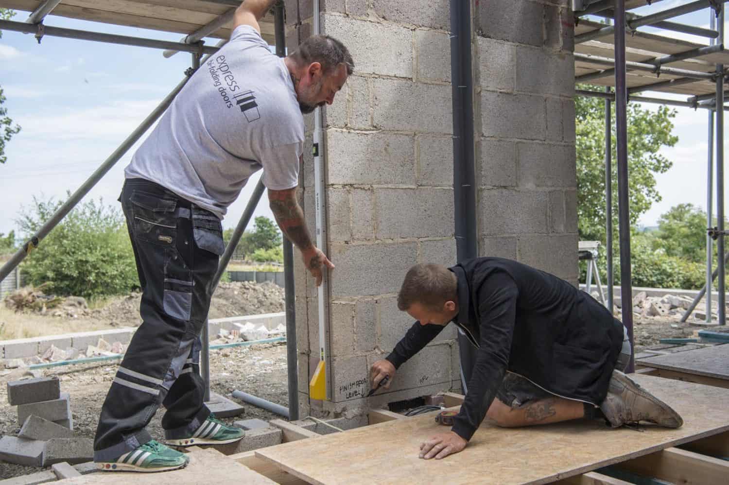 Two construction workers measure and mark a brick wall at a building site, possibly preparing for new front doors. One stands with a measuring tool while the other kneels, marking the wall. Scaffolding and construction materials are visible around them.