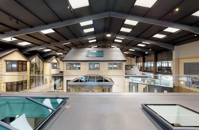 Interior view of a large showroom with high ceilings, skylights, and model house facades. The space features modern lighting, glass railings, bespoke glass solutions, and displays related to home improvement or construction.