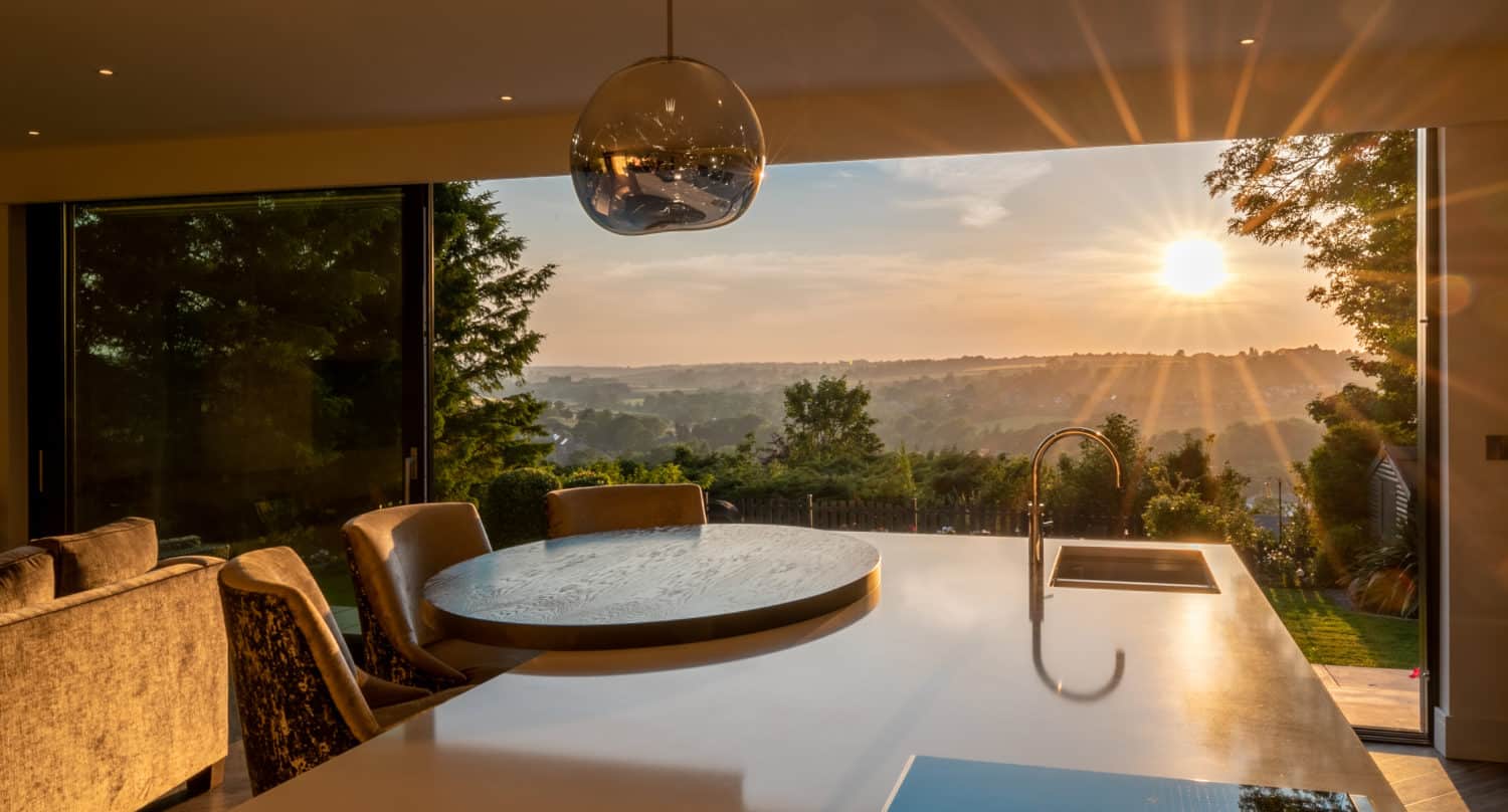 Modern kitchen with a large island, sink, and pendant light, opening to a patio through bifold doors with bespoke glass solutions. Sunlight streams in at sunset, filling the space with a warm glow and framing views of trees and hills.