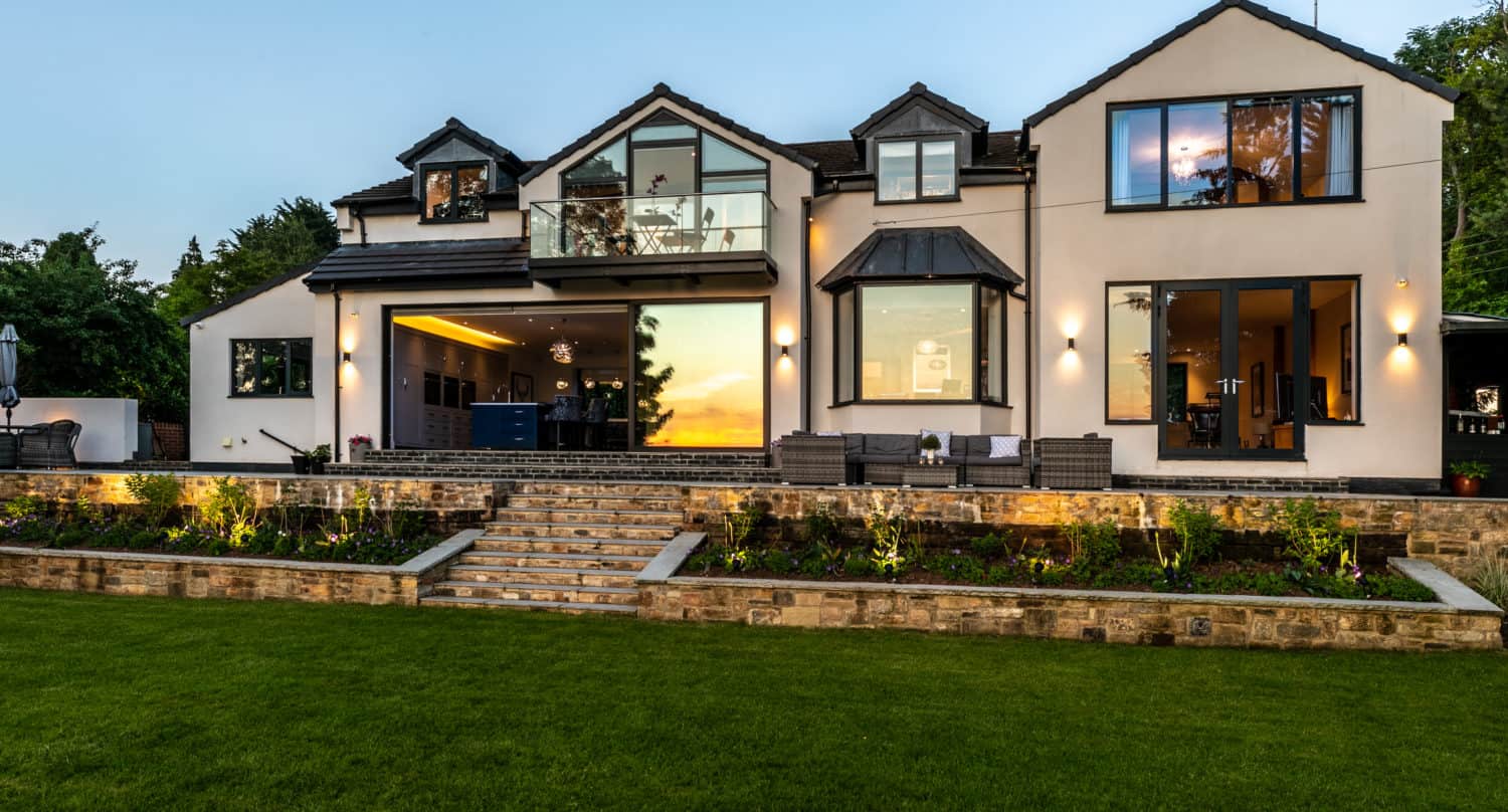Modern two-story house with large windows and exterior lights, featuring stylish bifold doors that open to a manicured lawn and stone steps leading to a landscaped garden; the sunset is reflected in the windows.