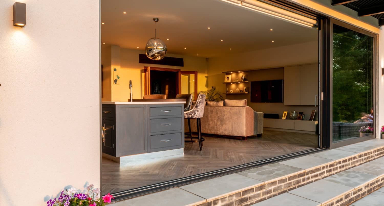 Open sliding bifold doors reveal a modern kitchen and living room with contemporary furniture, a kitchen island, and a hanging light fixture. Potted flowers sit outside on the stone patio.