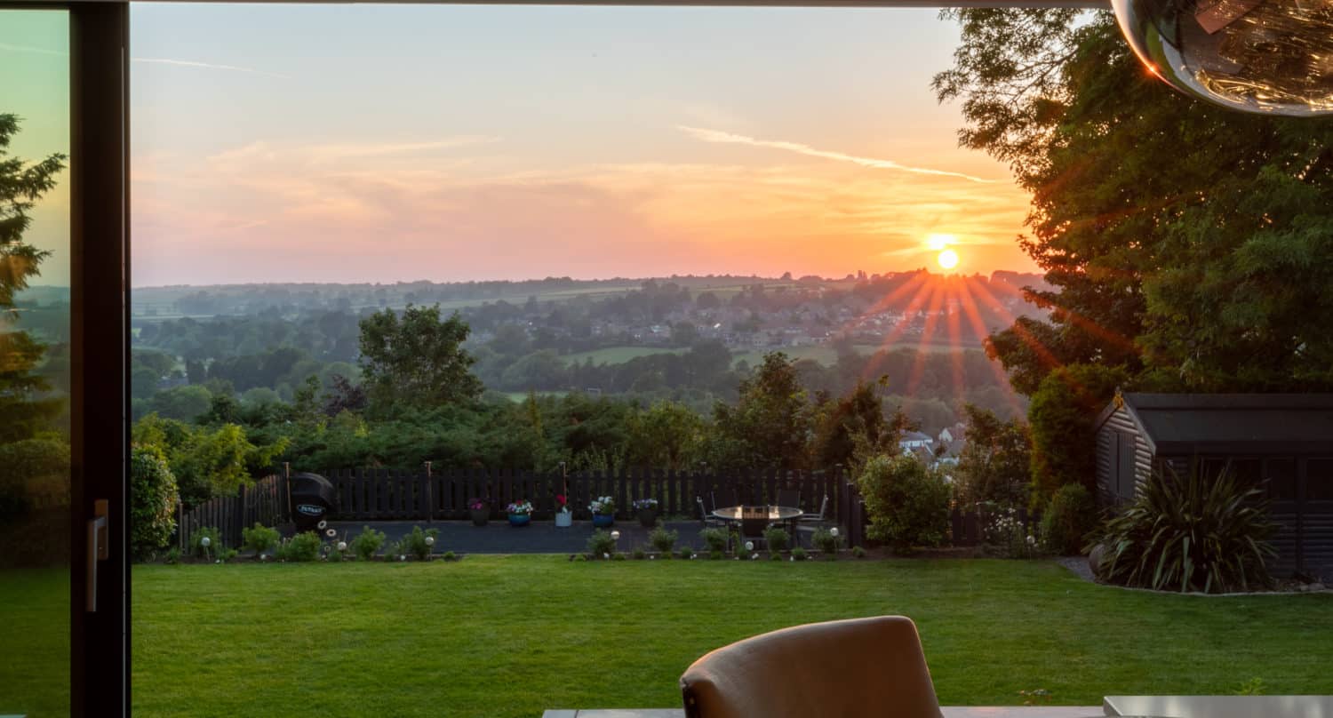 A view from a modern home’s dining area, looking out through sliding doors onto a green garden and a distant landscape at sunset, with the sun low on the horizon and soft orange light filling the sky.