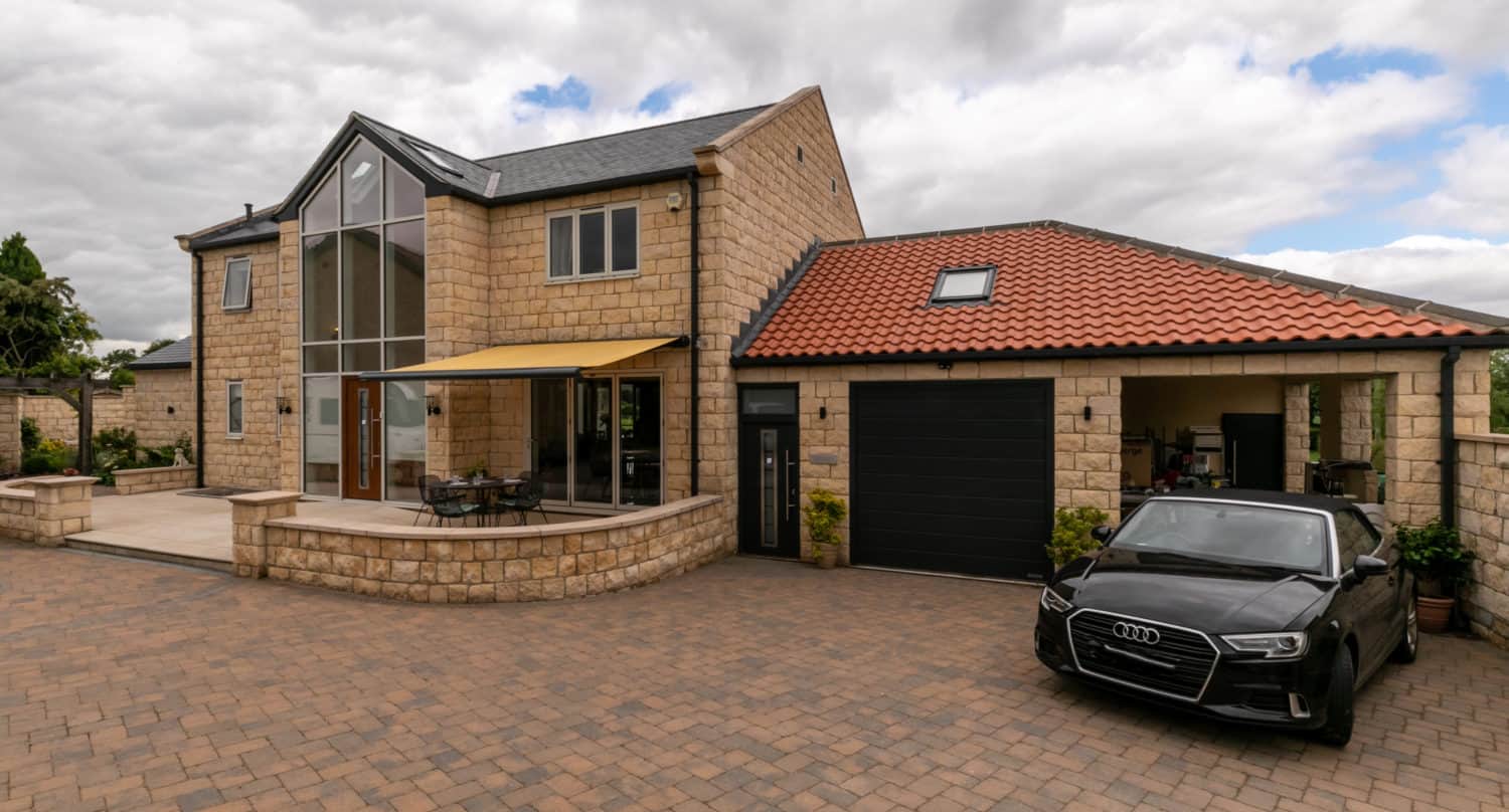Modern stone house with large windows, bespoke glass solutions for the front doors, and a red-tiled roof. A black Audi is parked in the driveway, and there is a covered patio area with outdoor furniture. The sky is mostly cloudy.