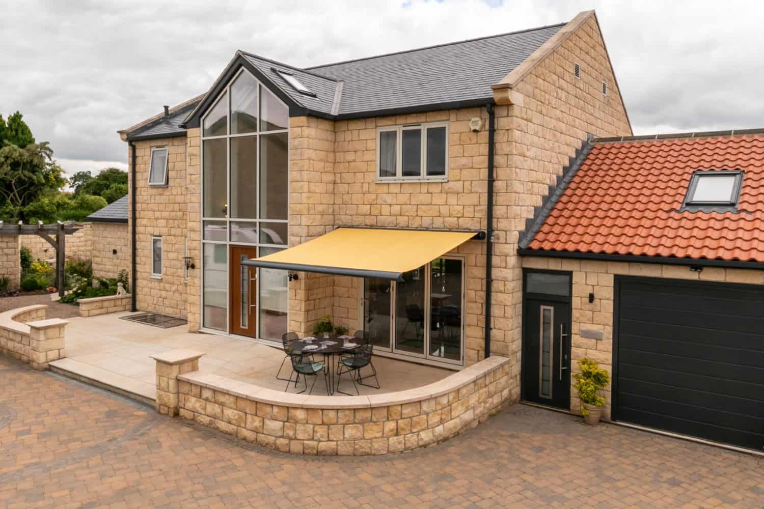 Modern two-story stone house with large glass windows and sleek sliding doors, a yellow retractable awning covering a patio dining area, and an attached garage with a black door. The yard is paved and landscaped with greenery.
