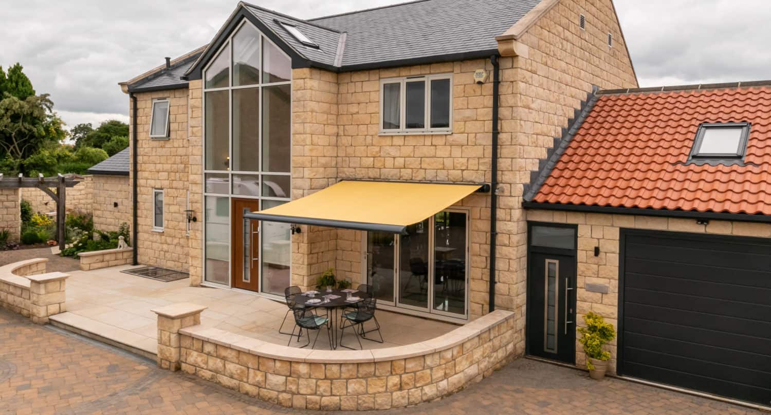 Modern two-story stone house with large glass windows and sleek sliding doors, a yellow retractable awning covering a patio dining area, and an attached garage with a black door. The yard is paved and landscaped with greenery.