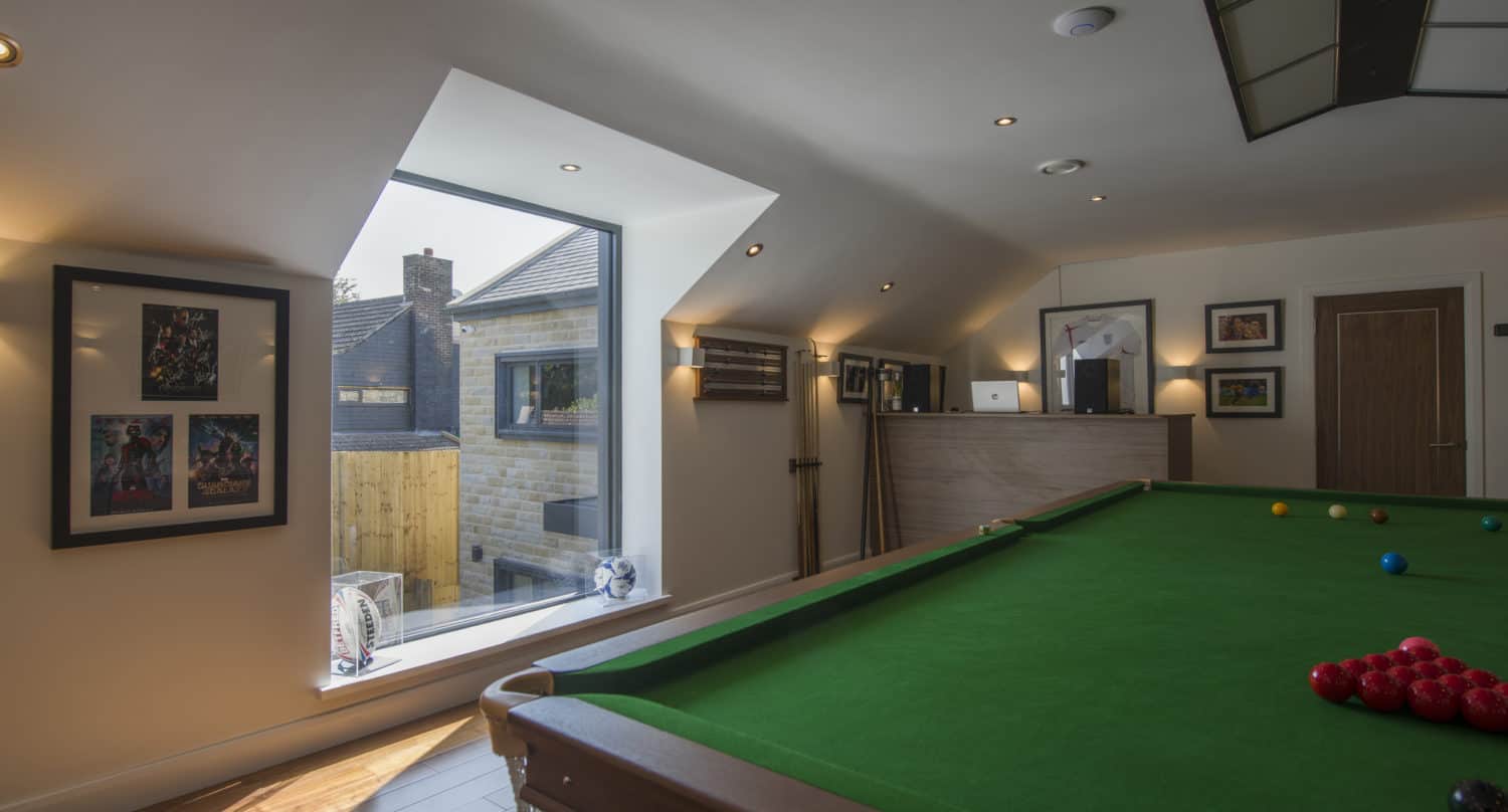A well-lit attic game room with a green snooker table, framed pictures on white walls, wooden floors, and a sloped ceiling featuring spotlights and bespoke glass solutions that let in natural light from a large window overlooking neighboring buildings.