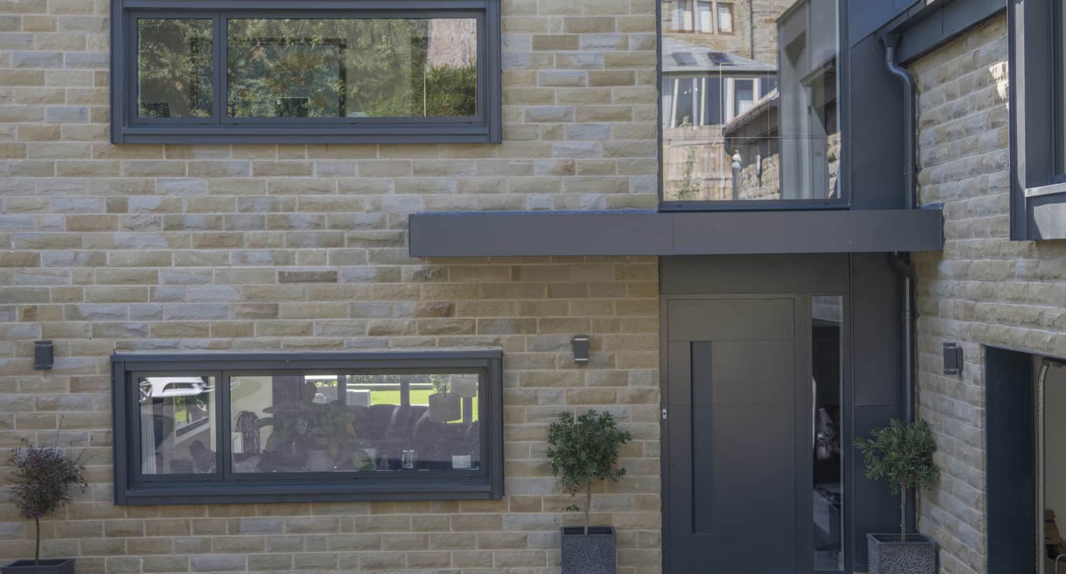 Modern house facade with light brown brickwork, large rectangular windows with gray frames, a gray front door, sliding doors opening to the garden, two potted plants by the entrance, and a black paved pathway in front.