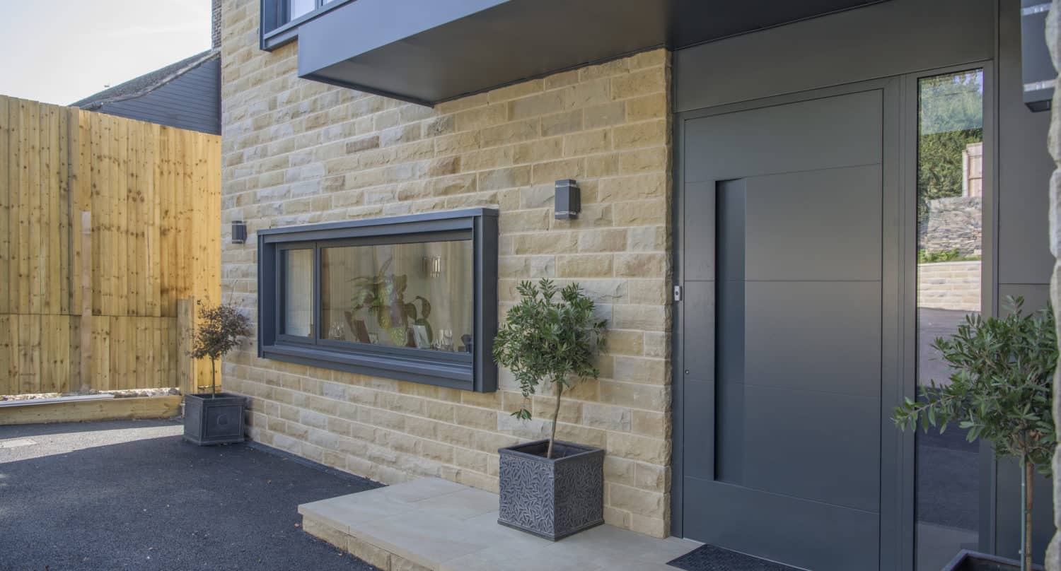 Modern house entrance with a gray front door, stone exterior, large horizontal window featuring bespoke glass solutions, and potted plants on a small stone porch, next to a wooden fence on a paved driveway.