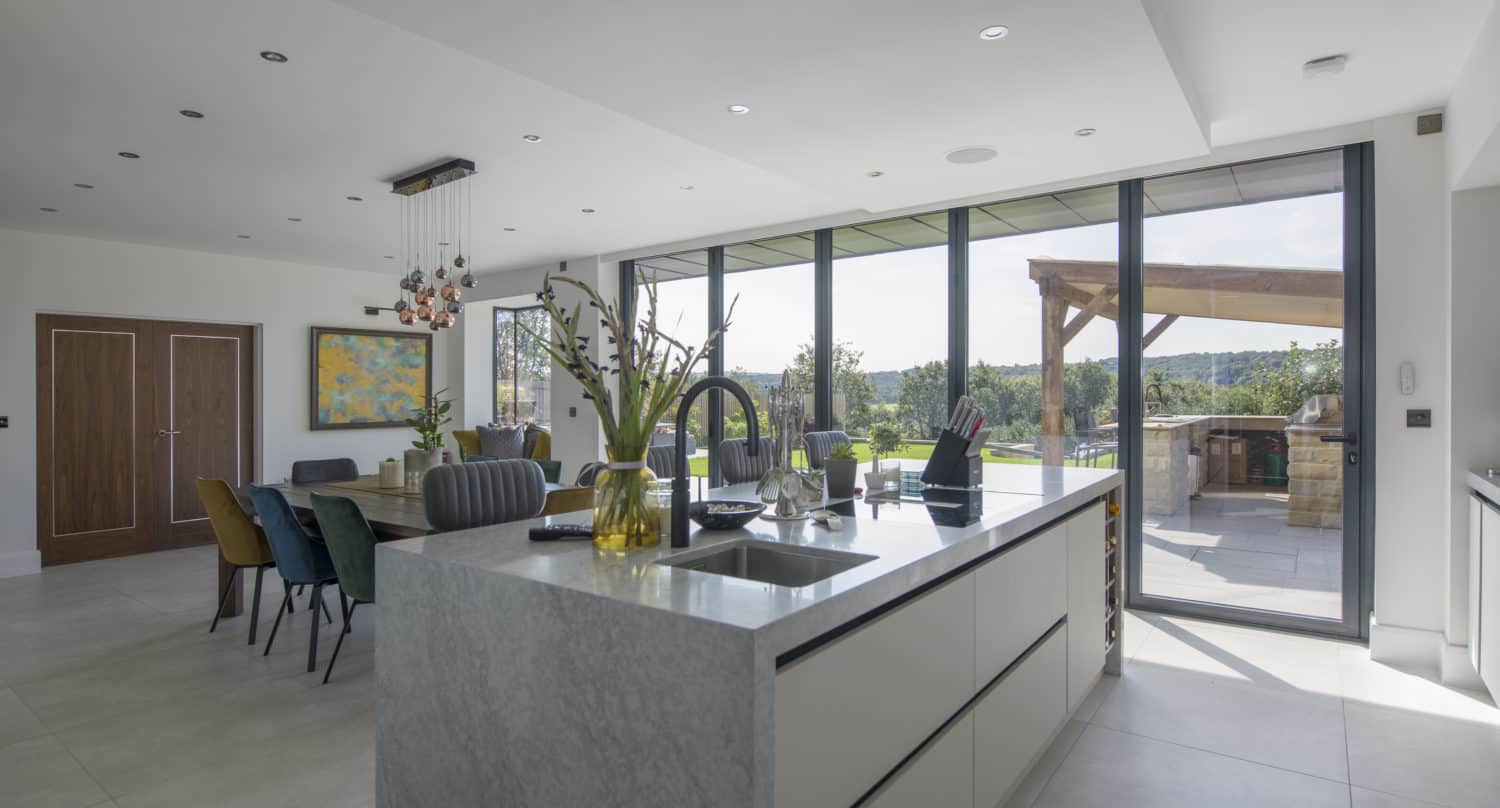 A modern, spacious kitchen with a large island, dining area, and floor-to-ceiling sliding doors opening to a sunny outdoor patio and garden. Bright natural light fills the space, decorated with plants and colorful chairs.
