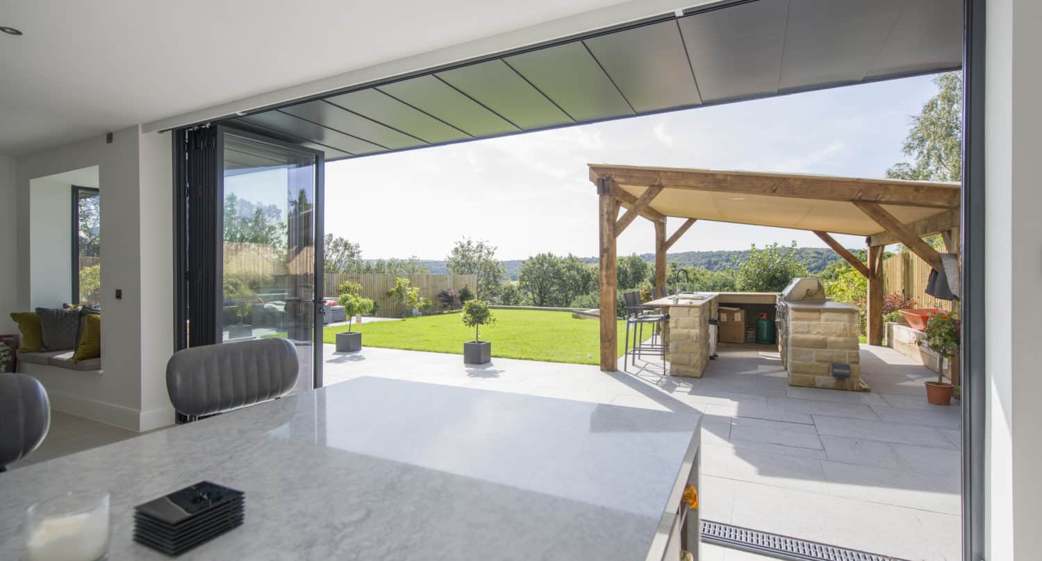 Modern kitchen with marble countertop opens to a spacious patio through bifold doors; outdoor wooden pergola with seating and a built-in grill are visible under a sunny sky.