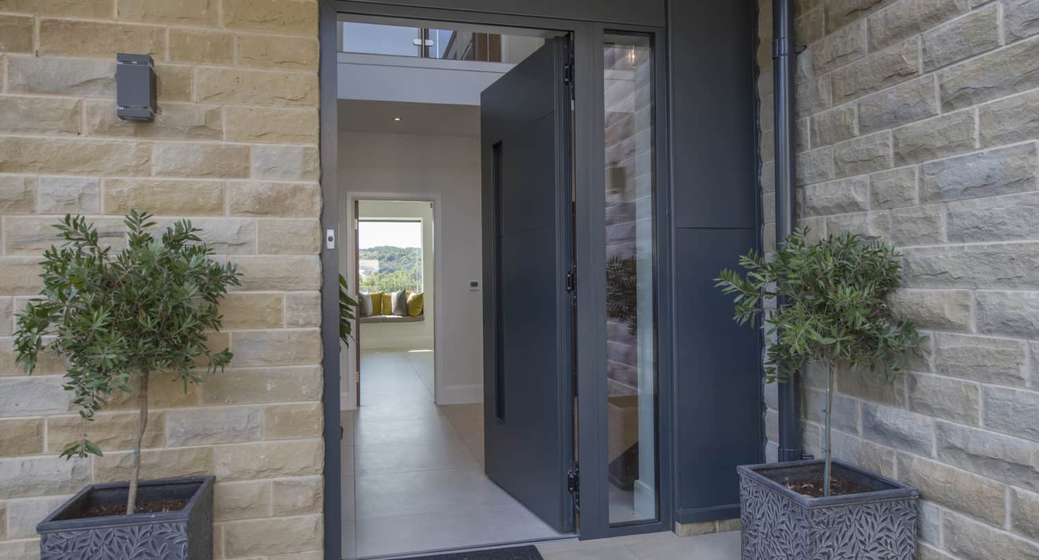 A modern front door is open, revealing a bright interior with a view of the living area and greenery outside. Two potted plants flank the entrance, while tan stone bricks and stylish front doors complete the inviting exterior.