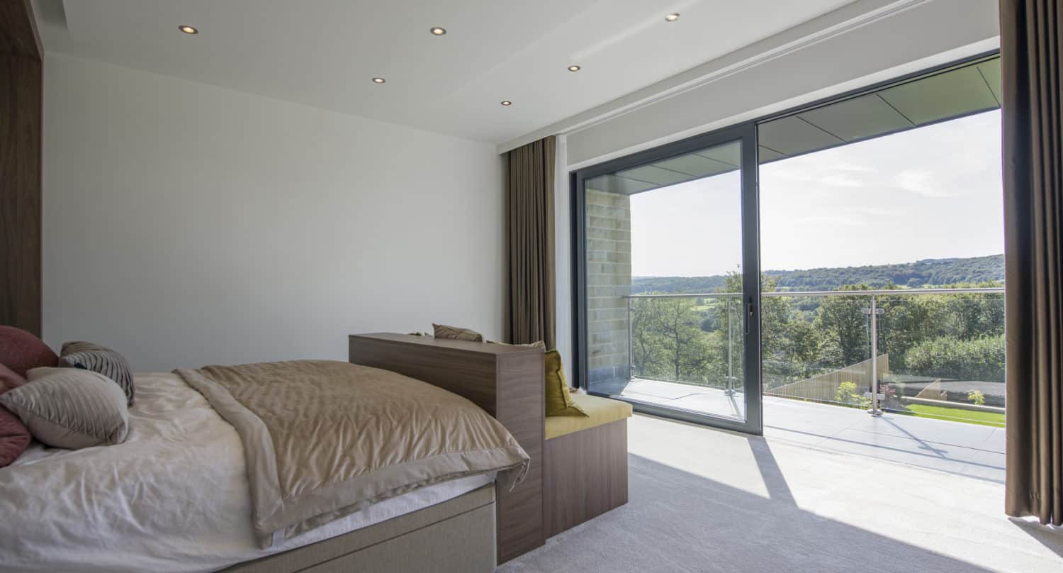 Modern bedroom with a neatly made bed, large bespoke glass sliding doors opening to a balcony with a scenic view of greenery and hills, and recessed ceiling lights.