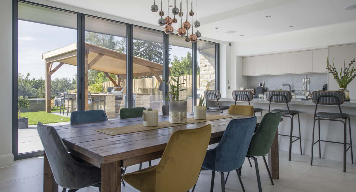 Modern dining and kitchen area with a large wooden table, colorful upholstered chairs, pendant lights, and bespoke glass solutions framing the view of a garden patio through floor-to-ceiling glass doors.