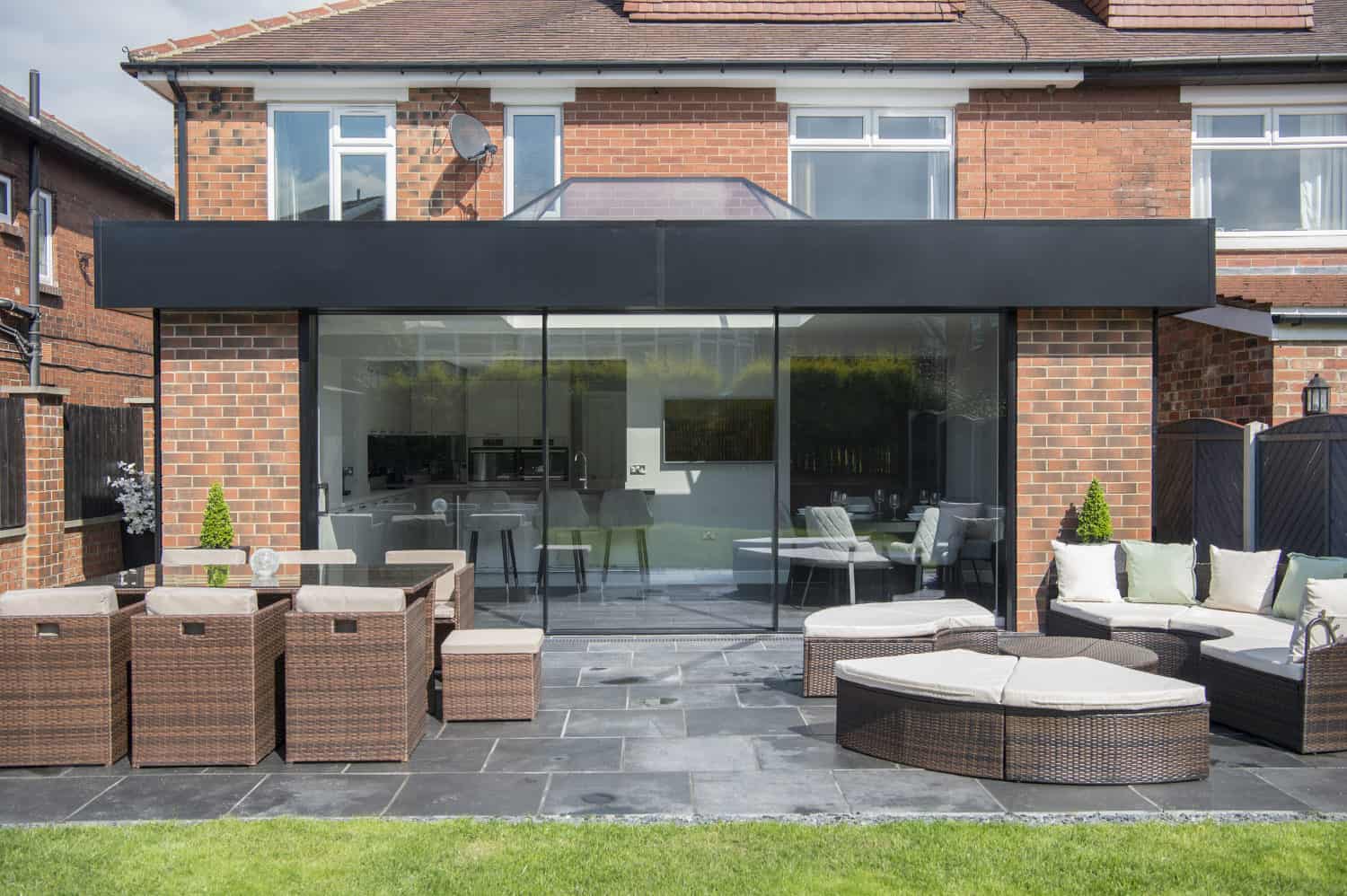 Modern brick house with large black-framed sliding doors opening to a patio with wicker furniture and a lawn. The patio features tables, chairs, white cushions for outdoor seating, and partial glass roofing for added style.