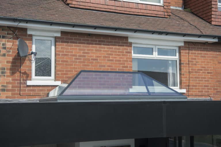 A glass pyramid-shaped skylight, part of the bespoke glass solutions, is set into the flat roof of a brick house extension, with two white-framed windows and a satellite dish visible on the main house wall above.
