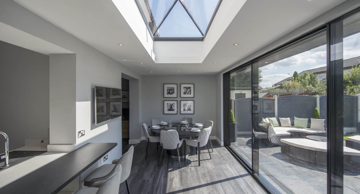Modern dining area with glass roofing, skylight, glass sliding doors opening to a patio with outdoor seating, gray walls, and minimalist décor. Dining table set for six, bar seating at a counter, and natural light filling the space.