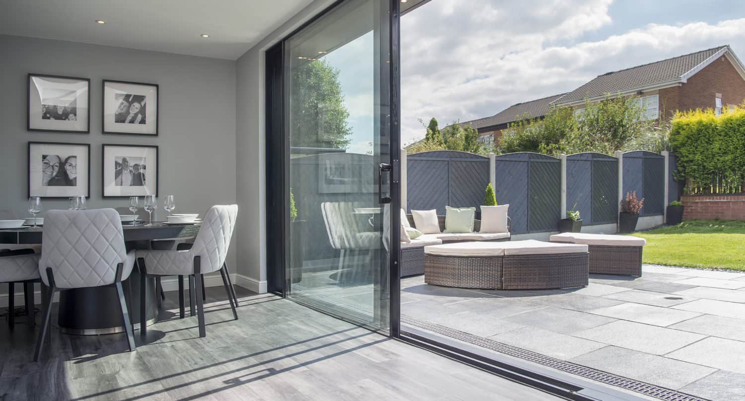 Modern open-plan dining area with a table and four chairs next to large bifold doors leading to a sunny patio with outdoor seating, surrounded by a privacy fence and greenery under a partly cloudy sky.