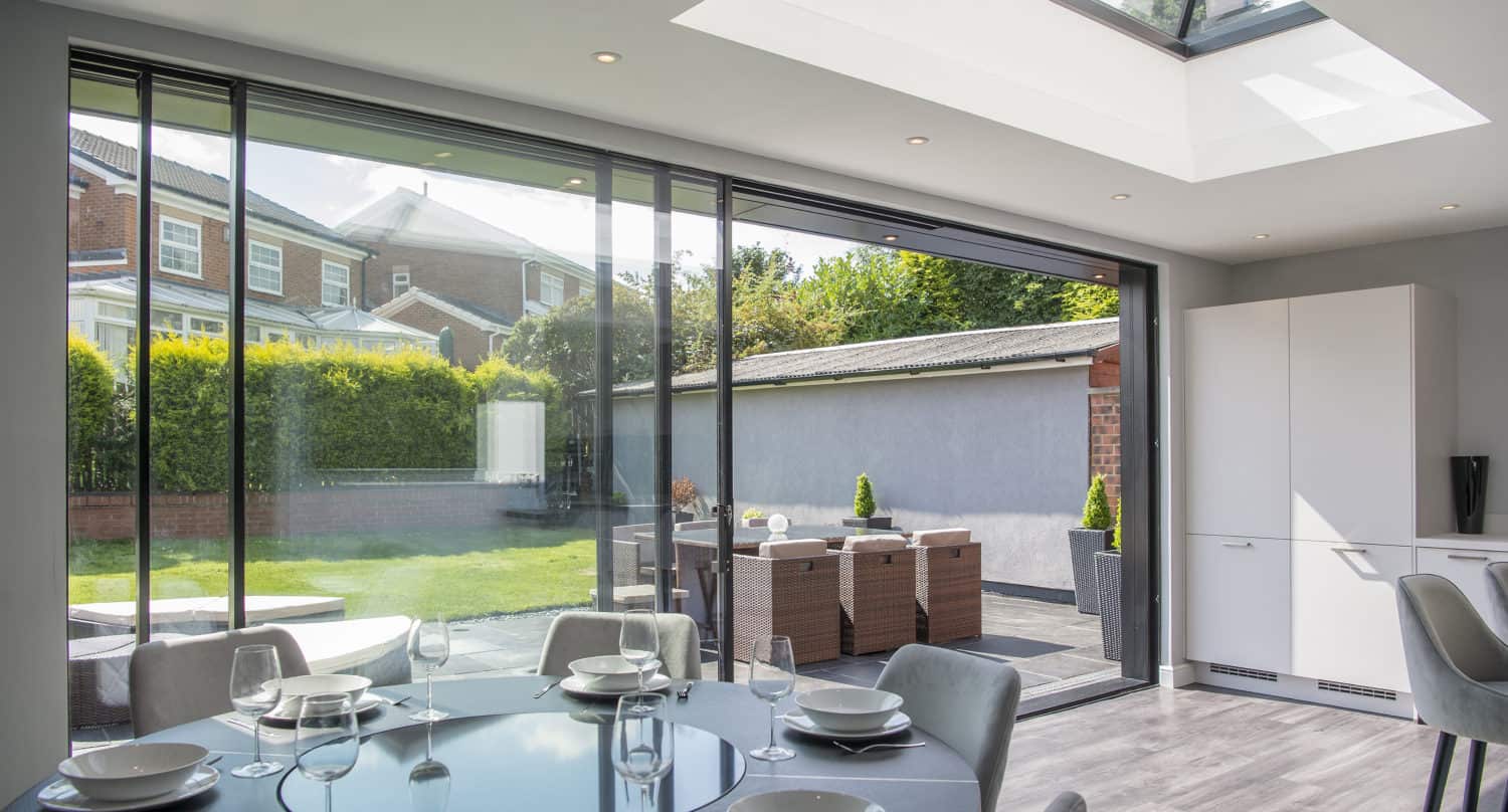 Modern dining area with a glass table and chairs, open-plan kitchen, large bifold doors, and skylight, overlooking a patio with outdoor dining set and a green backyard.