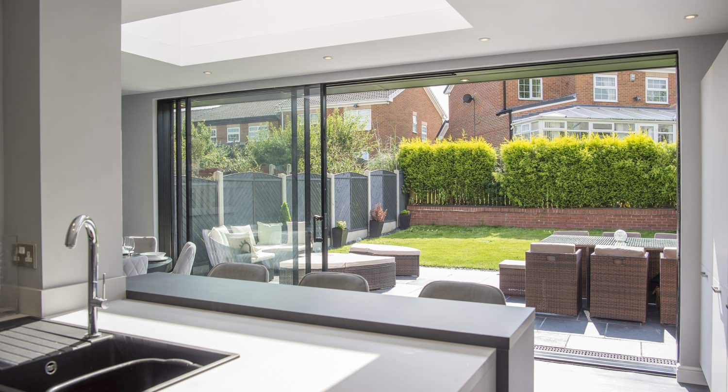 Modern kitchen with a sink in the foreground, looking out onto a patio with outdoor furniture and a well-kept garden, visible through large sliding doors and a skylight above featuring bespoke glass solutions.
