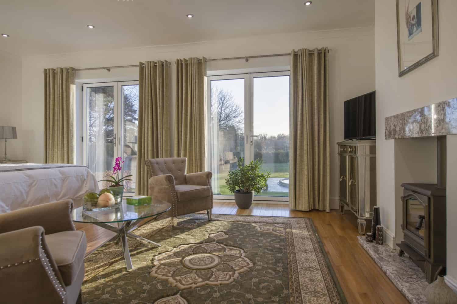 A bright, elegant living room with large windows, beige curtains, a patterned rug, modern furniture, a fireplace, TV, and green plants. Sunlight streams in through sliding doors with a beautiful view of trees outside.