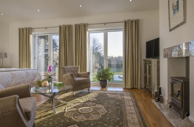 A bright, elegant living room with large windows, beige curtains, a patterned rug, modern furniture, a fireplace, TV, and green plants. Sunlight streams in through sliding doors with a beautiful view of trees outside.