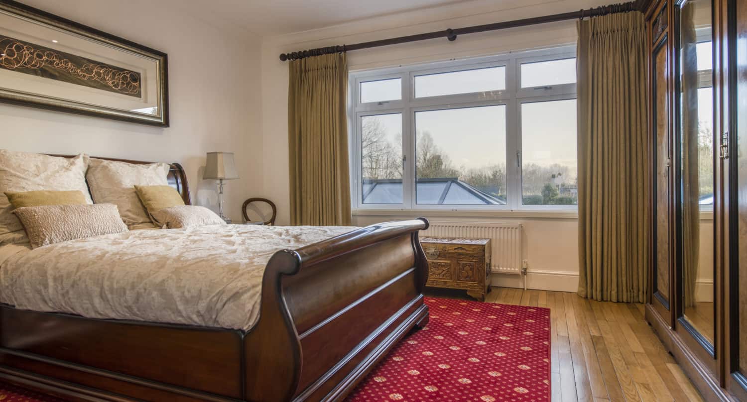 A cozy bedroom with a wooden sleigh bed, beige bedding, a red patterned carpet, large windows with sliding doors and curtains, a wooden chest, and a mirrored wardrobe on wooden floors.