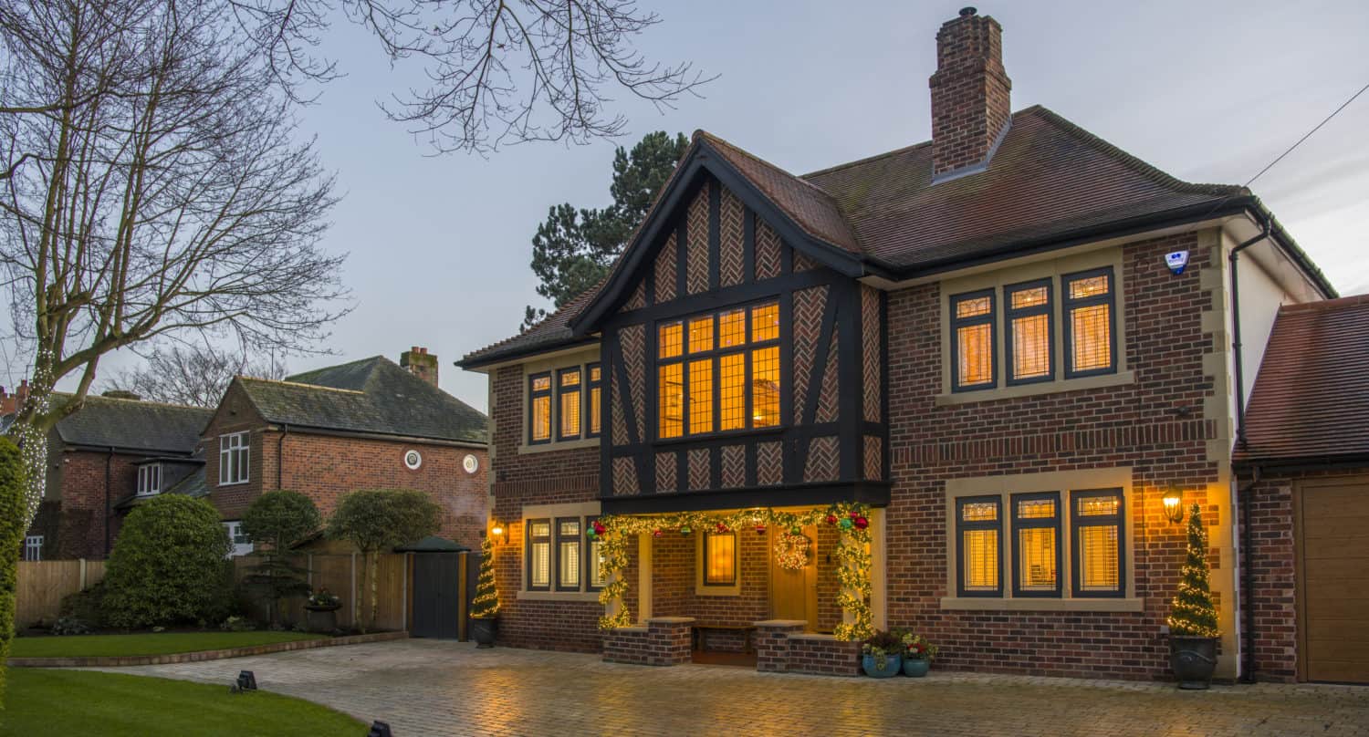 A two-story brick house with timber accents and glowing windows features elegant front doors and stands on a wet cobblestone driveway, surrounded by trees and bushes, with a neighboring house visible in the background at dusk.