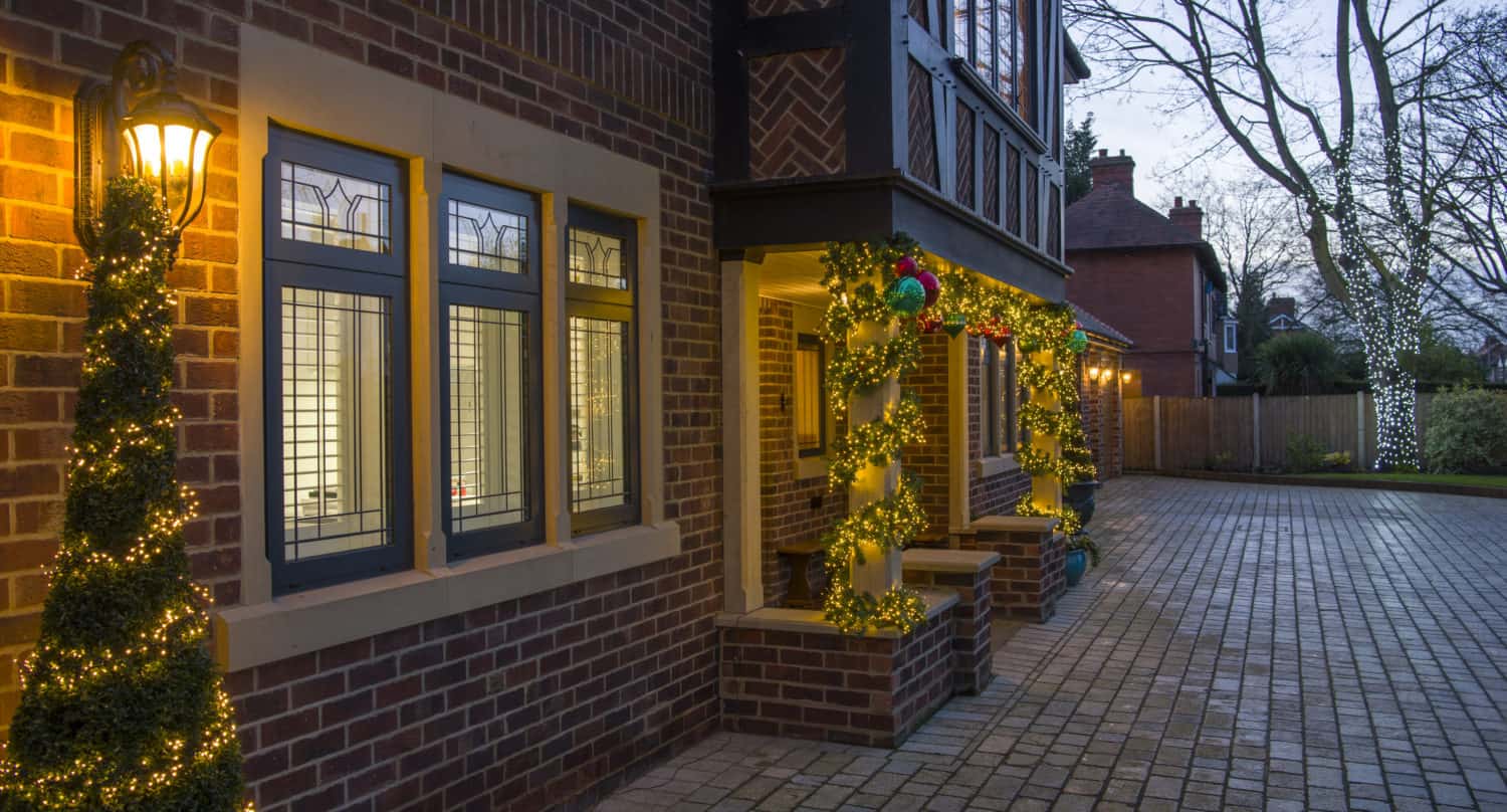 A brick house with bespoke glass solutions for the front doors, decorated with yellow string lights around the windows, door, and topiary at dusk. The brick driveway and trees in the yard are also wrapped with glowing lights.