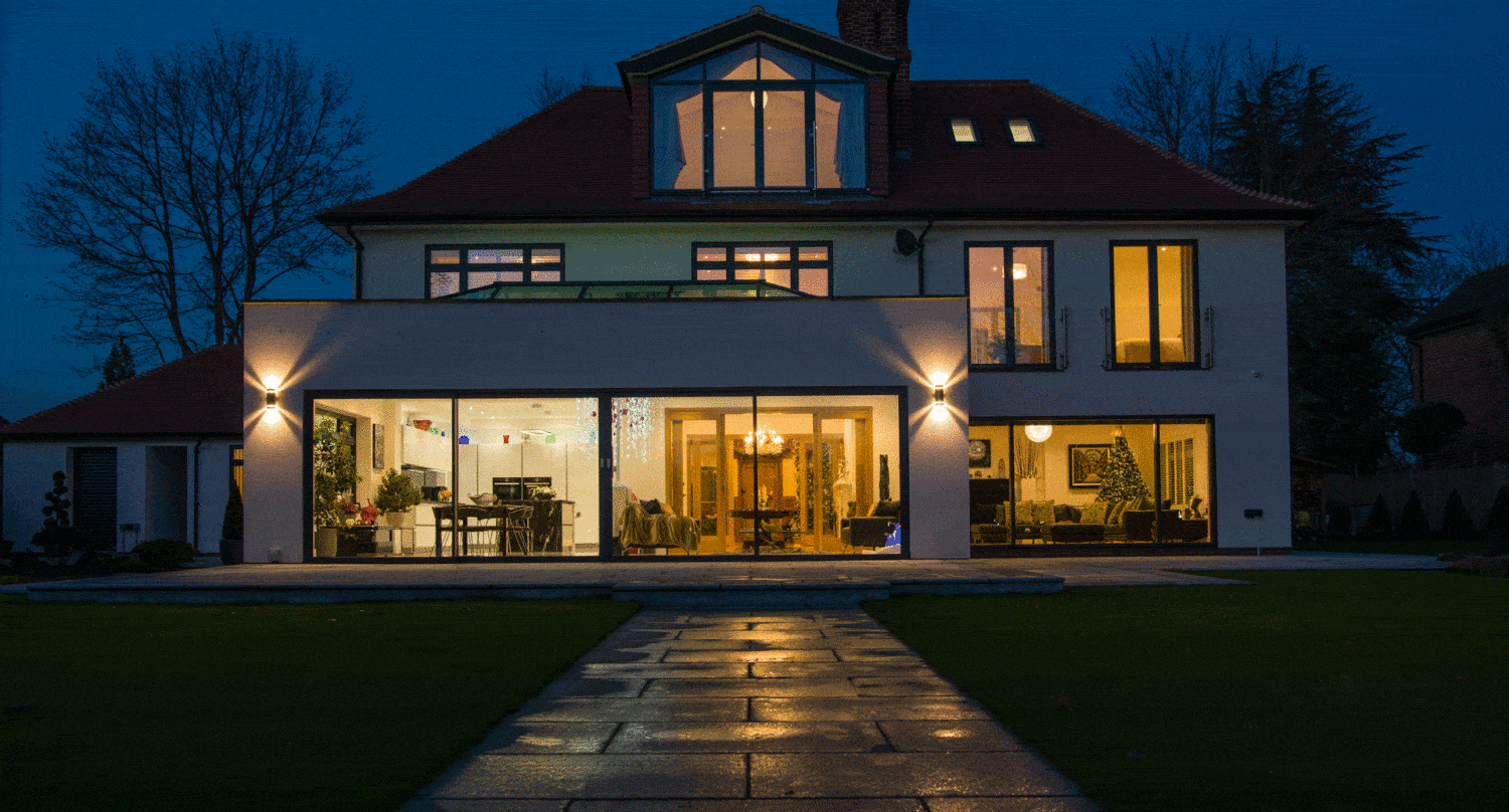 A modern two-story house with large windows and bespoke glass solutions is brightly lit from within at night. A wet stone pathway leads to stunning front doors, while the lawn and trees are dimly visible in the background.