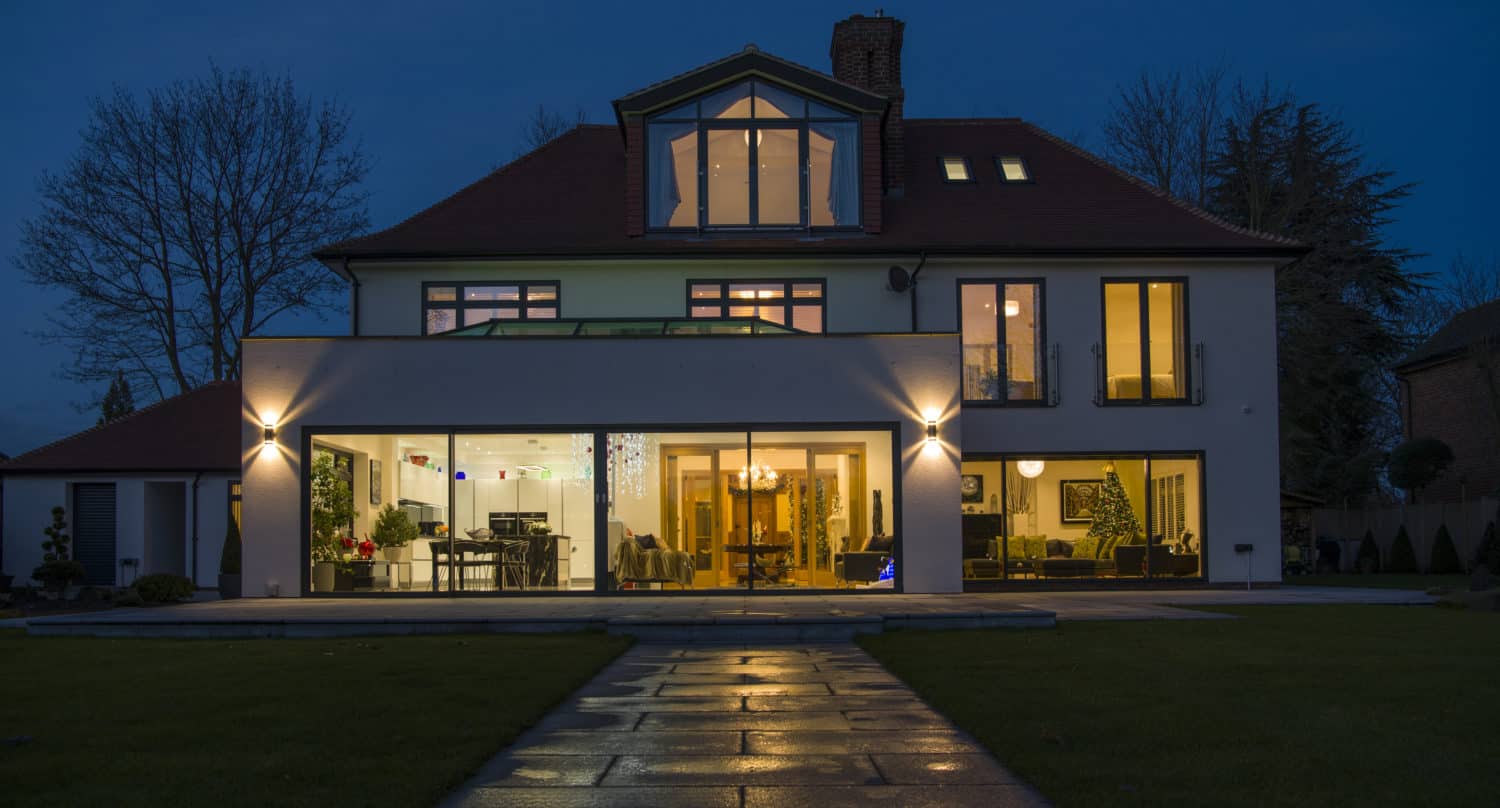 Modern two-story house at dusk with large illuminated windows, visible interior including furniture and plants, a stone pathway leading to the entrance, and sleek sliding doors. The sky is dark and trees are in the background.
