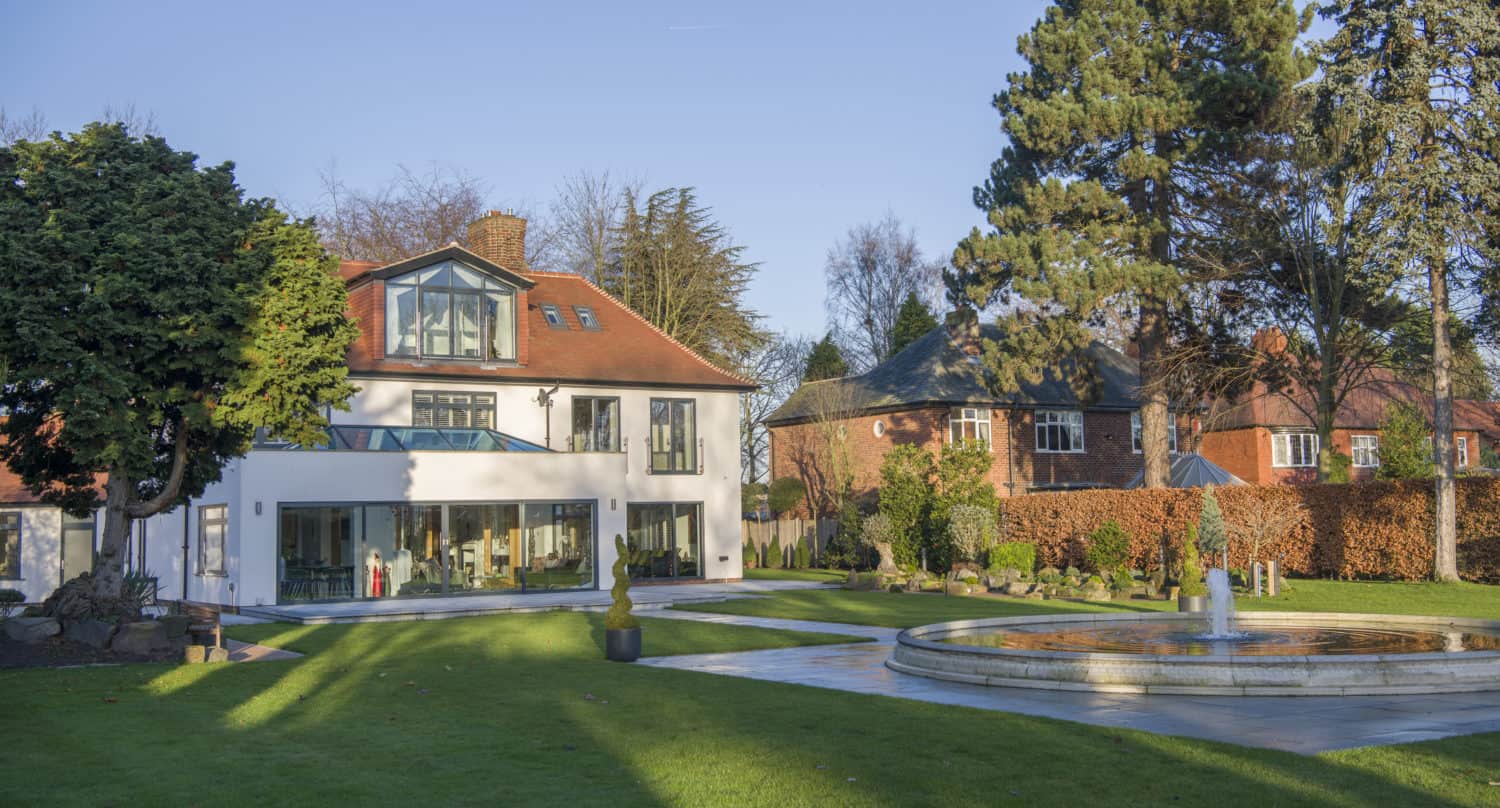 A large modern white house with big windows and bifold doors sits beside a spacious green lawn, mature trees, and a round stone fountain. Another brick house is visible in the background under a clear blue sky.