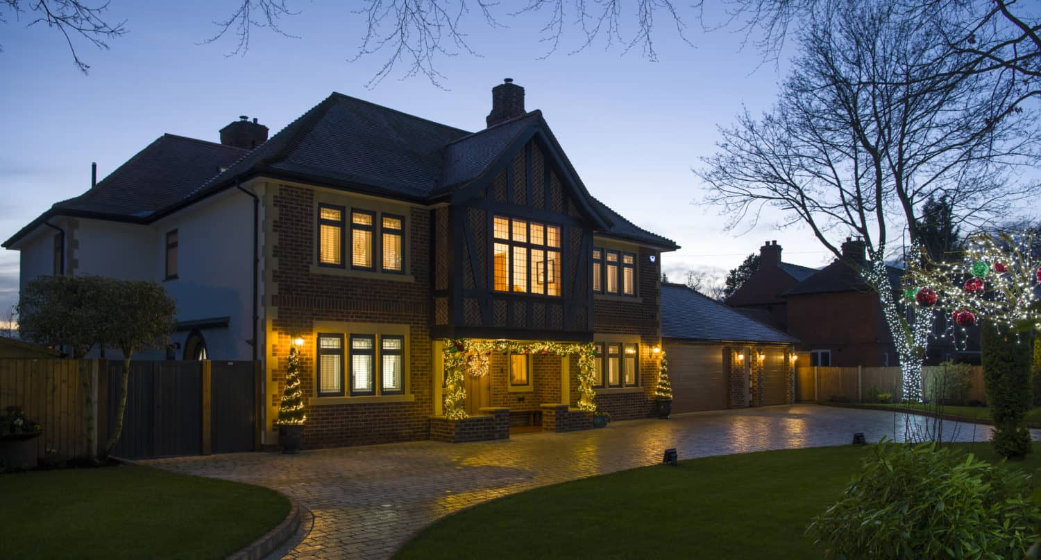 Two-story brick house with bespoke glass solutions like bifold doors and glass roofing, exterior lights glowing at dusk, a large driveway, and decorated trees with string lights and red ornaments. The sky is clear and the garden is neatly landscaped.