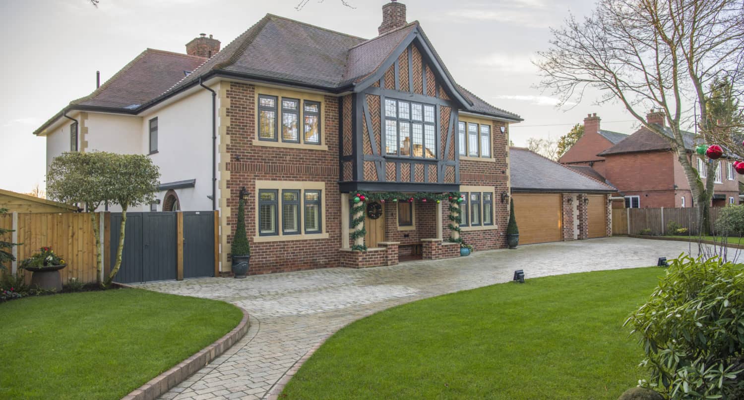 A large two-story house with brick and timber accents, bay windows, and elegant front doors. The well-manicured lawn features a curved stone pathway leading to the entrance, with trees and shrubs surrounding the property.