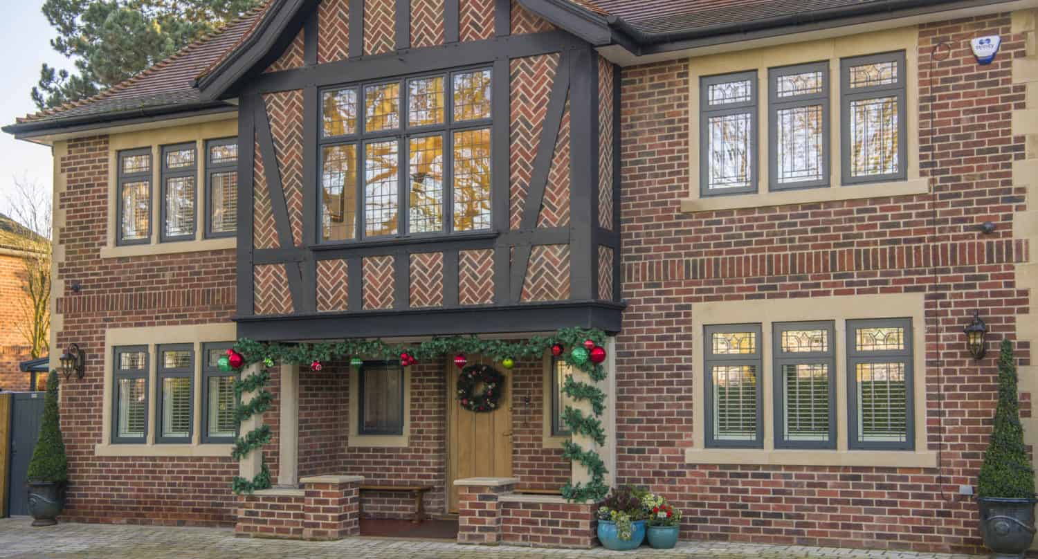 A two-story brick house with Tudor-style timber framing, decorated with green garland and red ornaments around the front doors. The windows have cream trim, and potted plants are placed by the entrance.