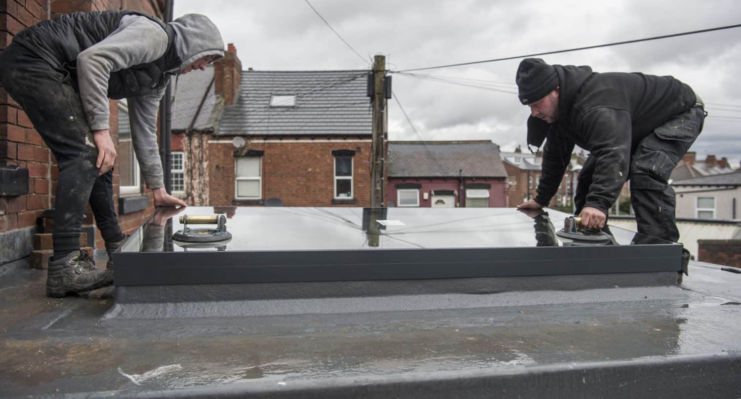 Two workers in hooded jackets and beanies install a large glass window panel on a flat rooftop, possibly as part of glass roofing, using suction handles, with brick houses and a cloudy sky in the background.