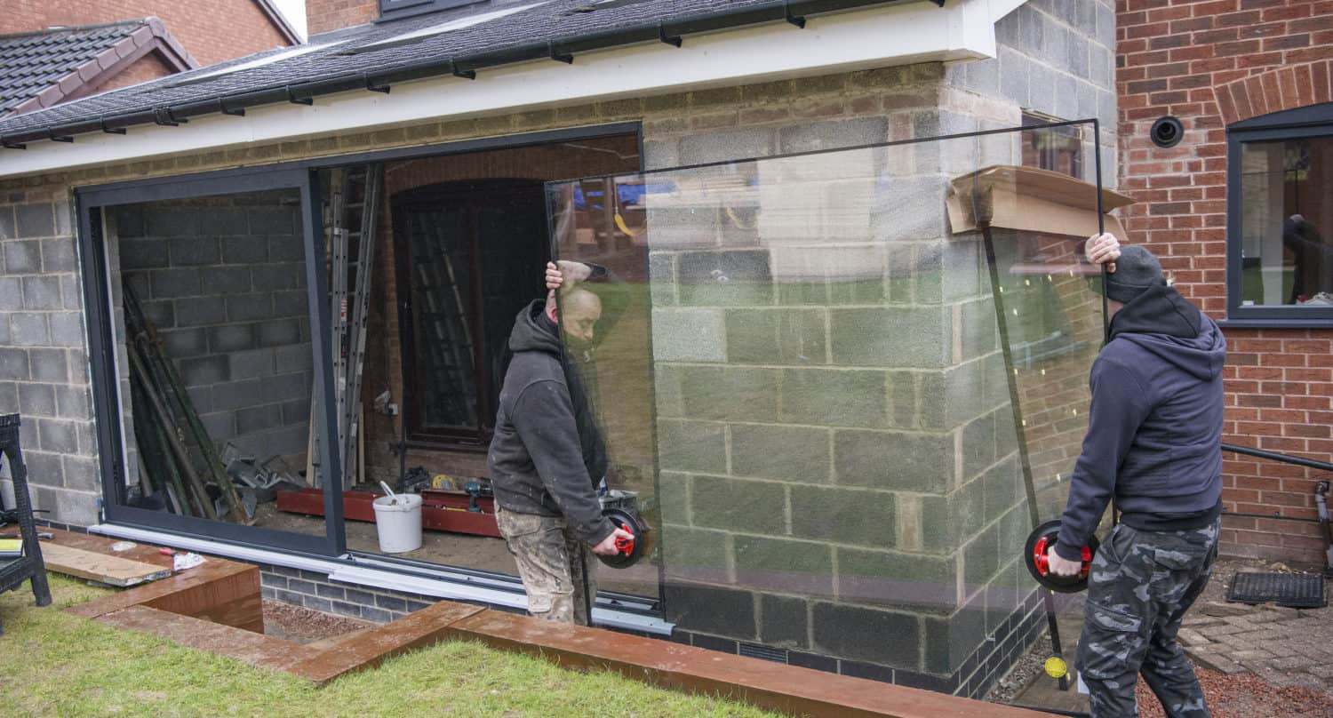 Two workers in hooded jackets install a large pane of glass in the opening of a partially constructed house extension, showcasing bespoke glass solutions. The extension features exposed block walls and construction materials inside.